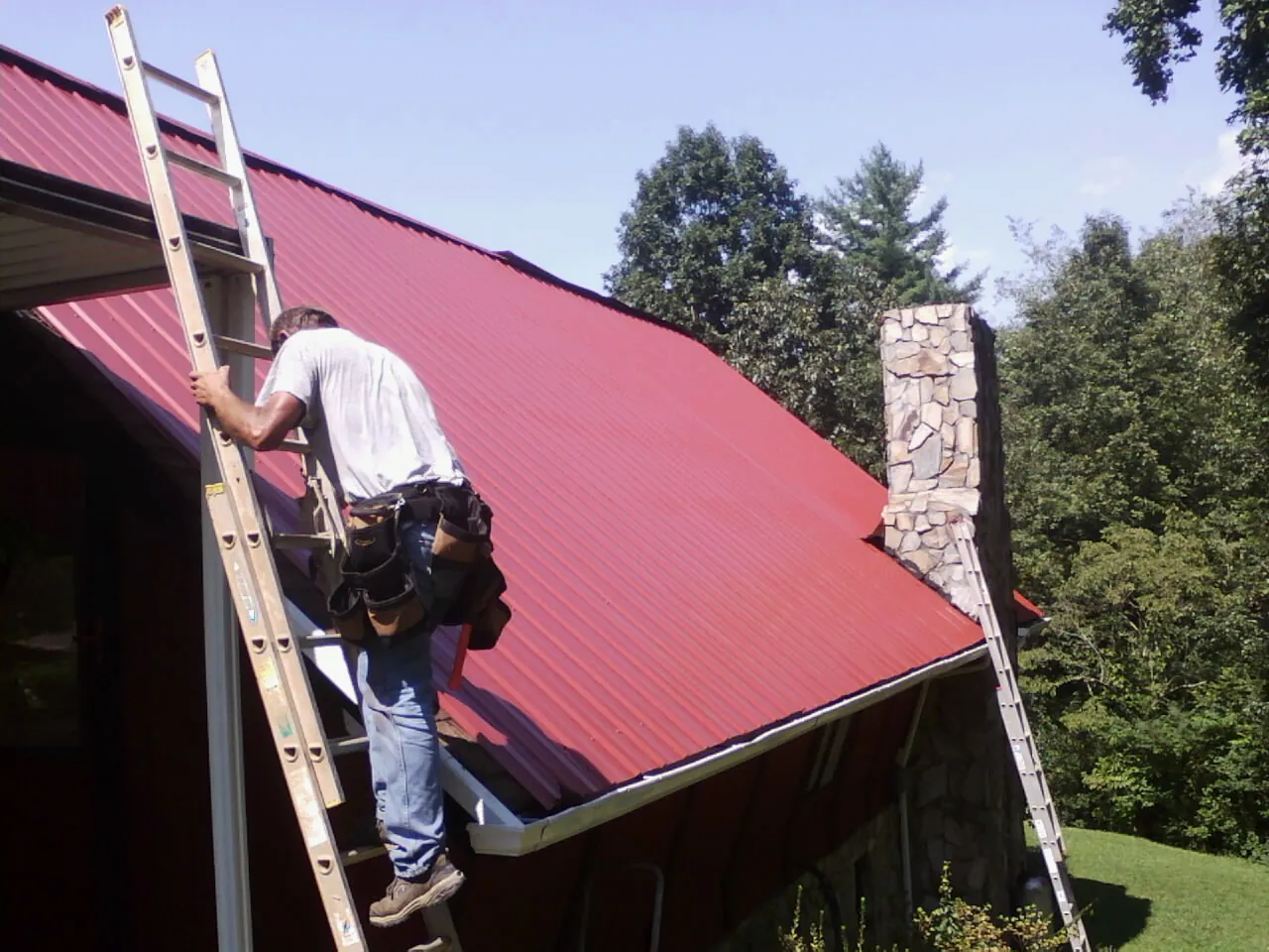 Roofer on a ladder beside a red metal roof and stone chimney — Soffit & Fascia Repair by Roofing Brothers in Montvale